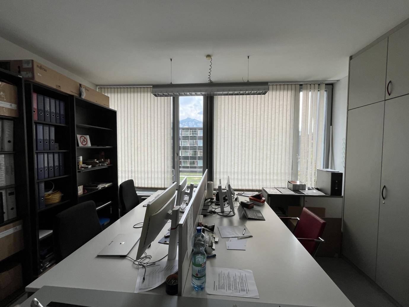 Großraumbüro mit mehreren Arbeitsplätzen, vertikalen Jalousien und Blick nach draußen.