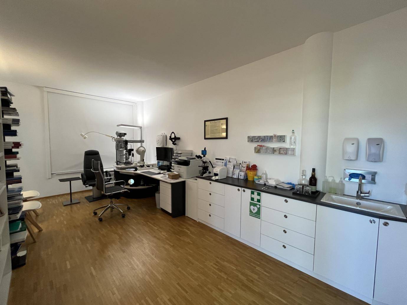 Praxisraum mit langer Arbeitsplatte, Waschbecken, medizinischen Geräten und Fenster mit Rollo.