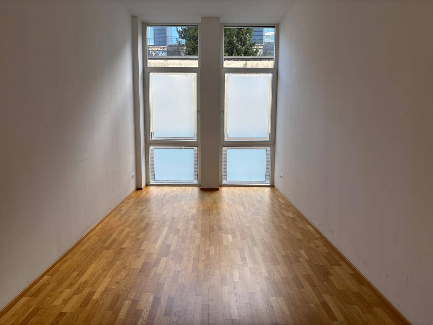 Leerer Raum mit hellem Holzfußboden und großen Fenstern, die viel Tageslicht hereinlassen.