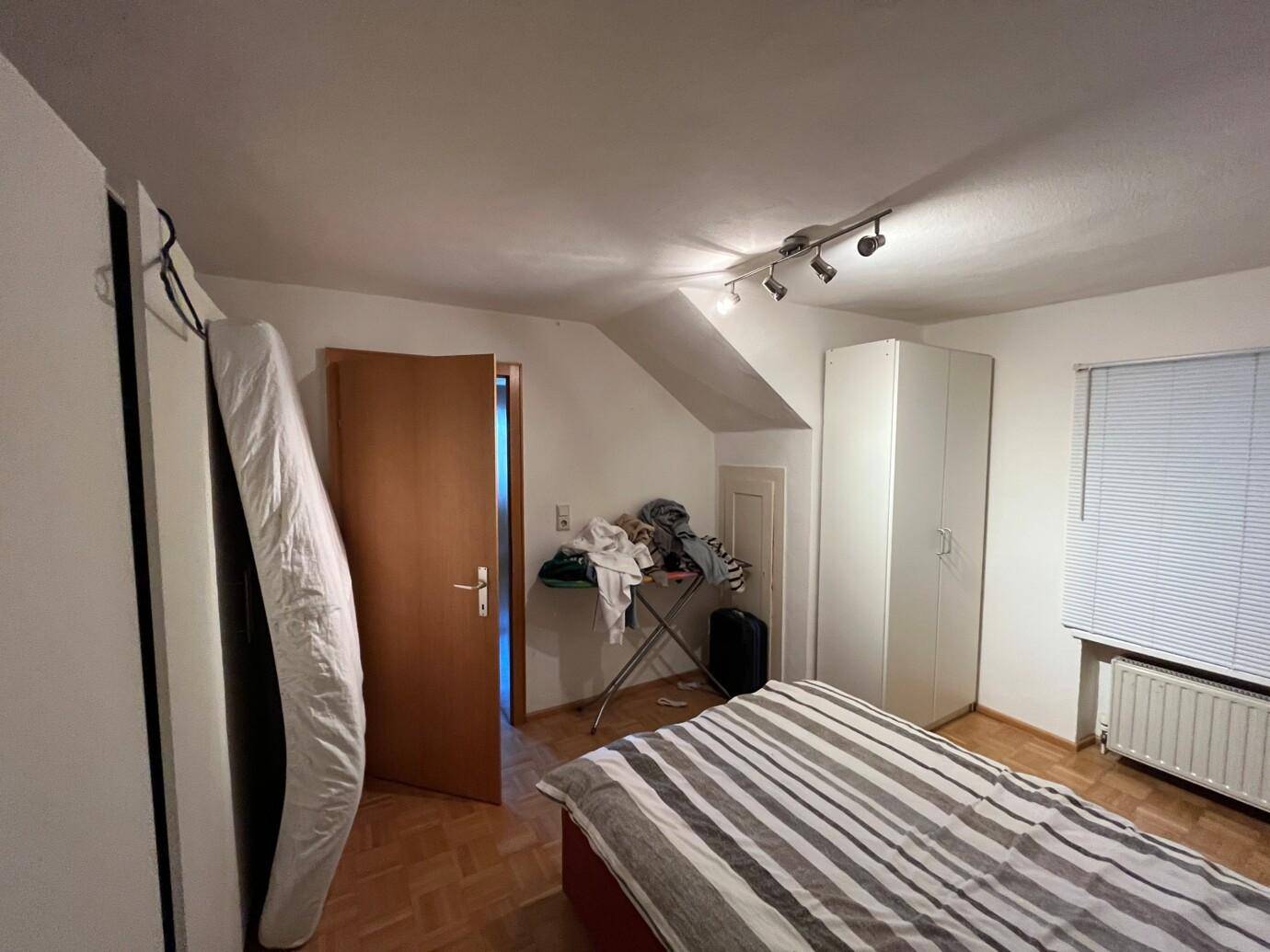 Unordentliches Schlafzimmer mit gestreiftem Bett, Bügelbrett voller Kleidung, Matratze an der Wand und Kleiderschrank.