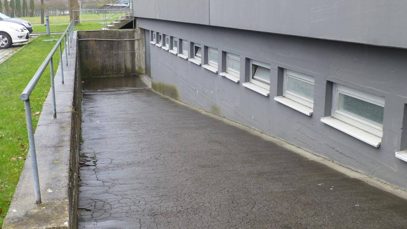 Außenansicht des Gebäudes mit kleinen Kellerfenstern und einem gepflasterten Zugangsweg entlang der Fassade.