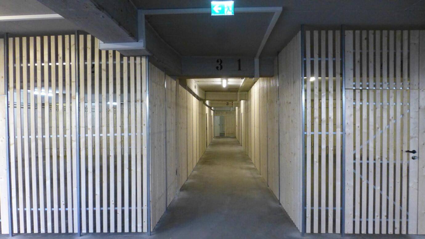 Langer Korridor mit Holzlattenwänden und Betonboden, führt zu Lagerräumen oder Parkplätzen im Untergeschoss.