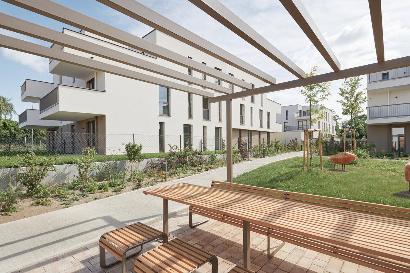 Gemeinschaftlicher Gartenbereich mit Pergola, Holztischen und Bänken zwischen modernen Gebäuden.