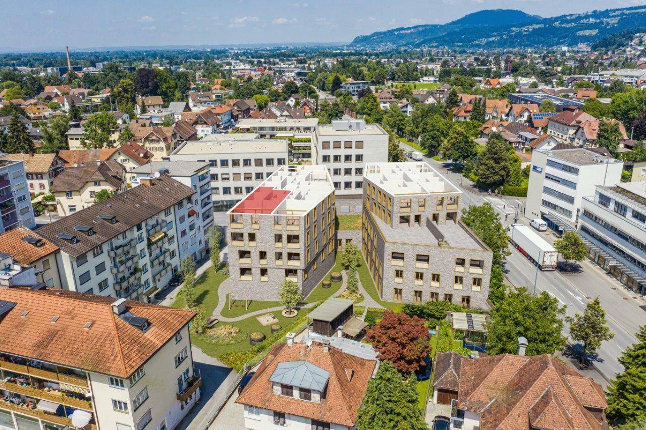 Luftaufnahme eines modernen Wohnkomplexes mit Neubauten, umgeben von bestehenden Gebäuden und Grünflächen, mit Bergen im Hintergrund.