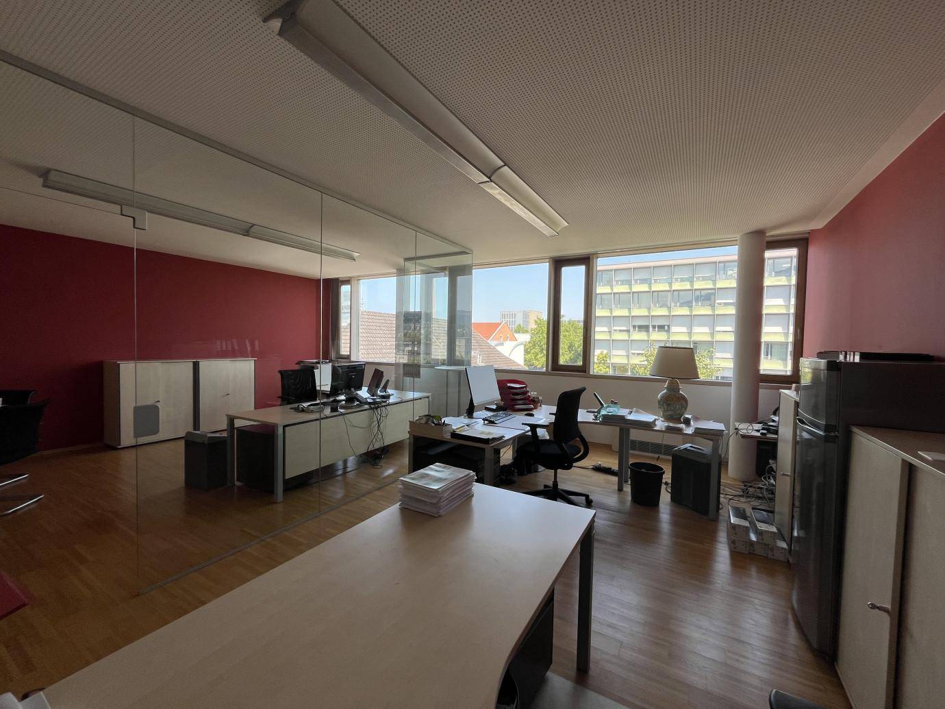 Helles Büro mit roten Wänden, großen Fenstern und mehreren Arbeitsplätzen mit Holzboden.