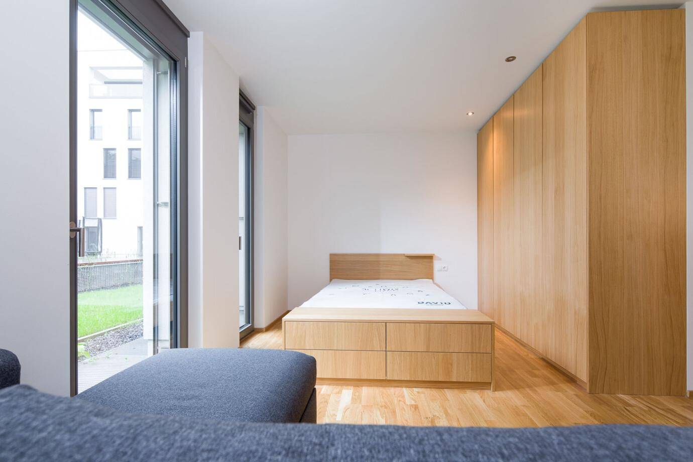Modernes Schlafzimmer mit Holzbett, integrierten Nachttischen, Holzkleiderschrank und Fenster mit Blick auf Grünfläche von Rhomberg Bau (David).
