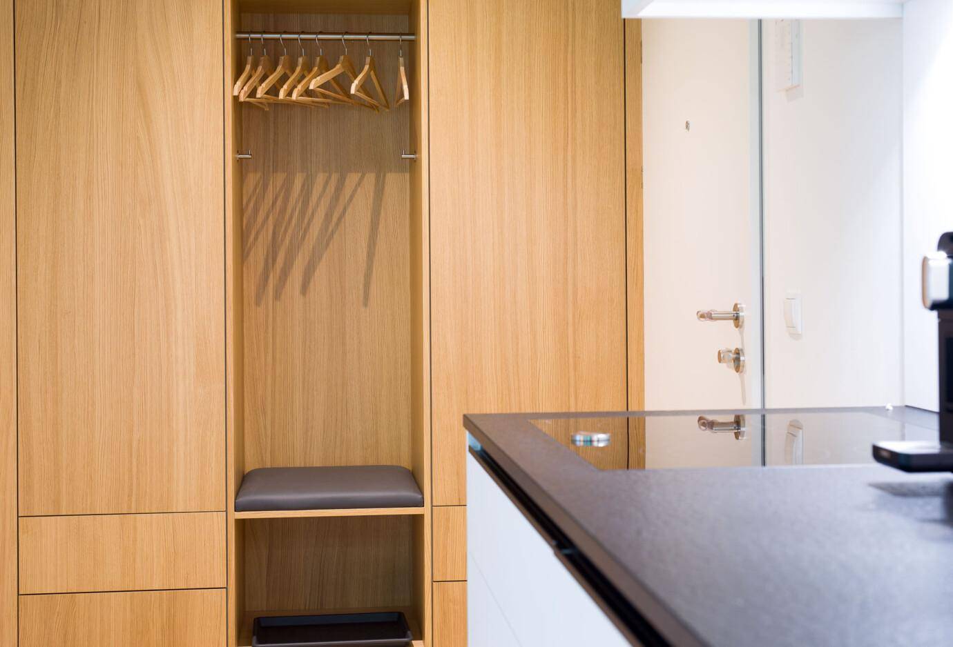 Holzschrank mit Kleiderstange, Bügeln und Sitzbank, daneben ein Spiegel und eine weiße Tür mit silbernen Griffen (David).