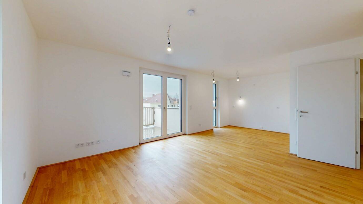Helles, leeres Wohnzimmer mit hellem Holzparkett und Zugang zu einem Balkon.