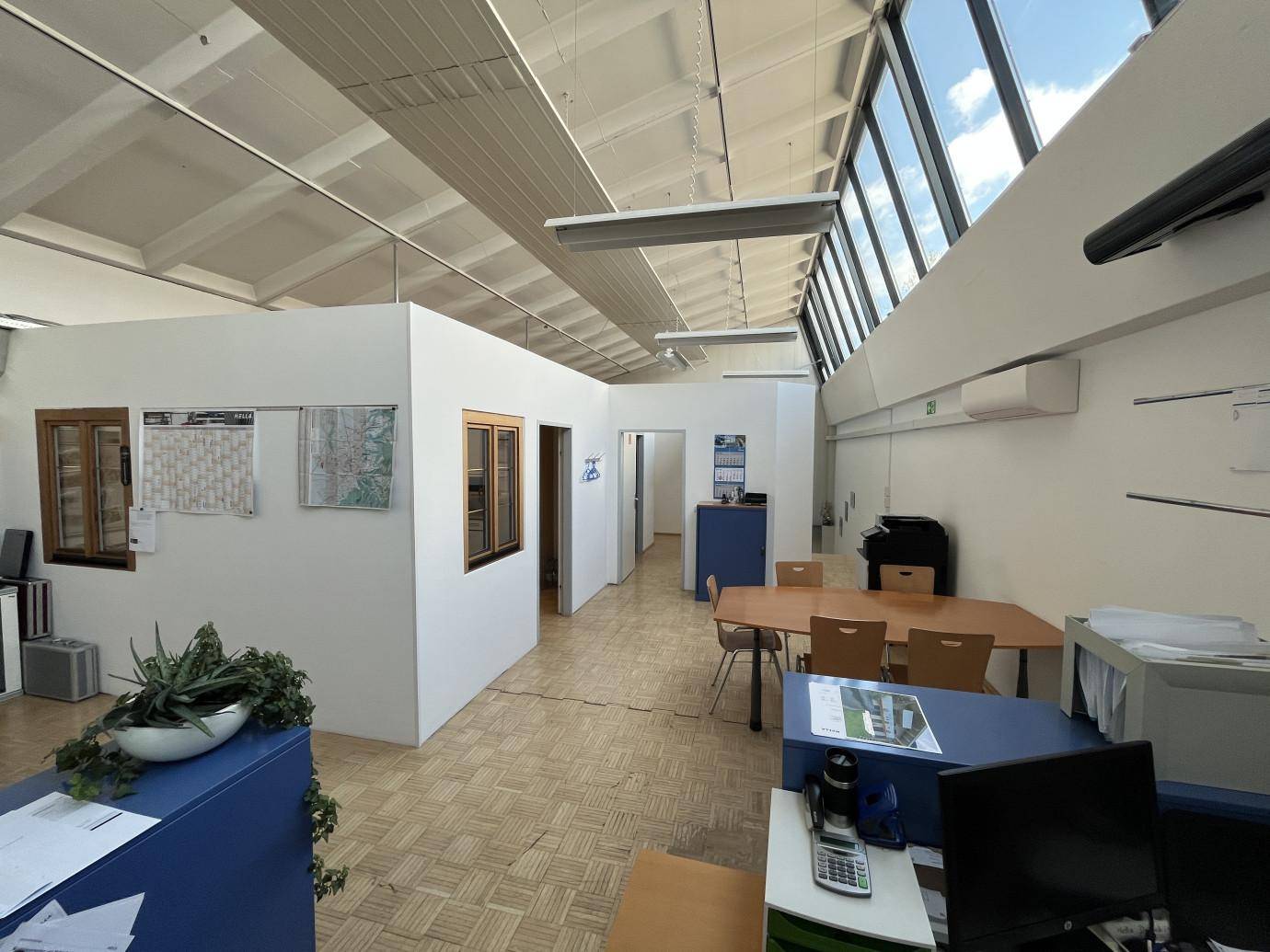 Offener Bürobereich mit Arbeitsplätzen, einem Flur und großen Fenstern, die viel Tageslicht hereinlassen.