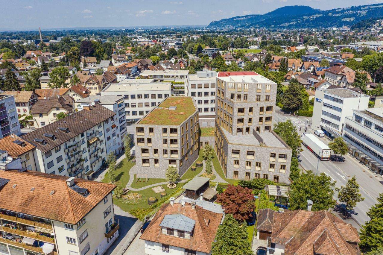 Luftaufnahme eines modernen Wohnkomplexes mit Gründächern und Spielplatz, eingebettet in eine städtische Umgebung mit grünen Hügeln im Hintergrund.
