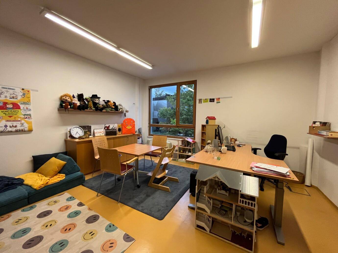Geräumiger Büro- oder Therapieraum mit Schreibtischen, Kindermöbeln und einem Fenster mit Blick ins Grüne.