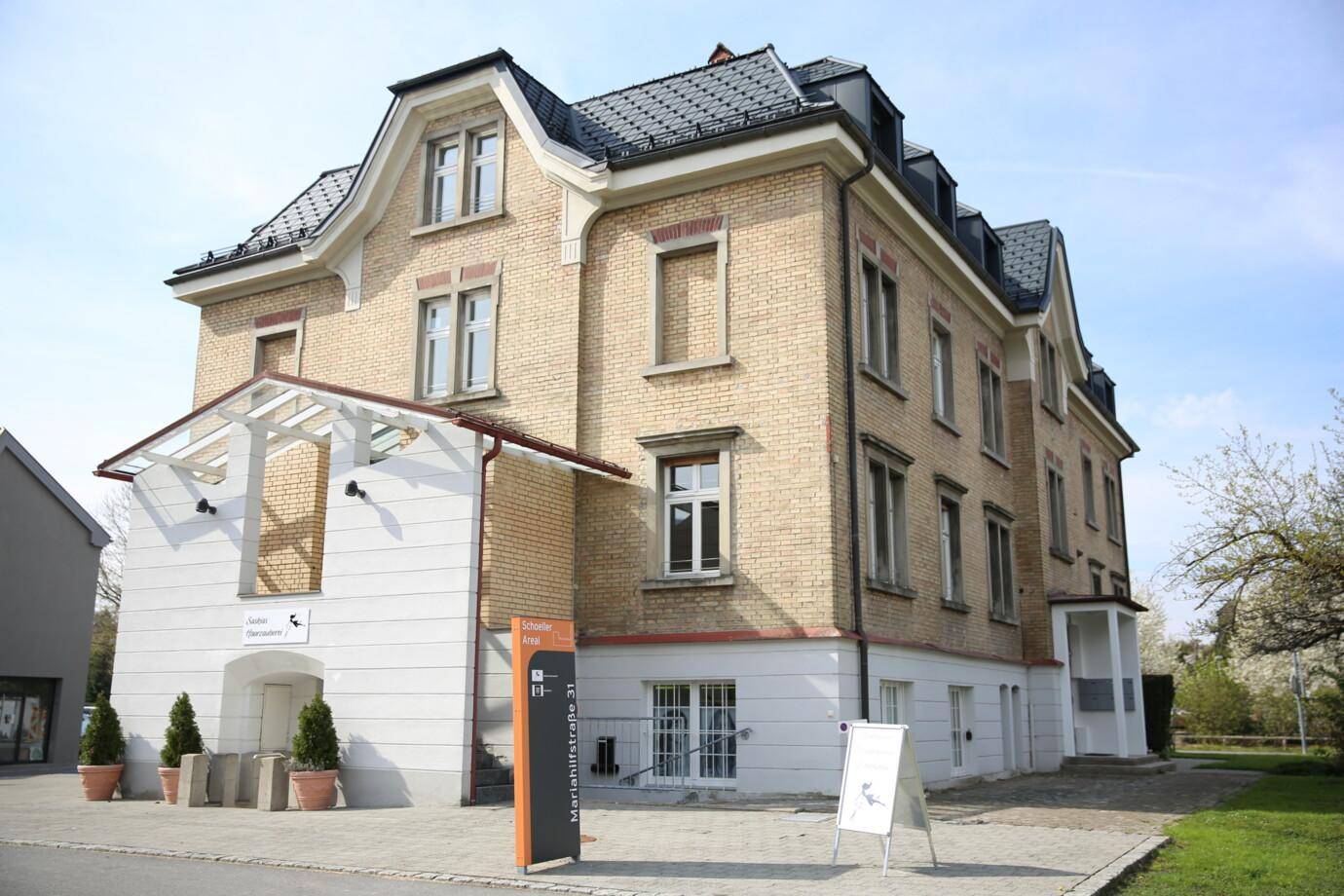 Historisches Backsteingebäude mit Mansarddach und modernen Anbauten in einer grünen Umgebung.