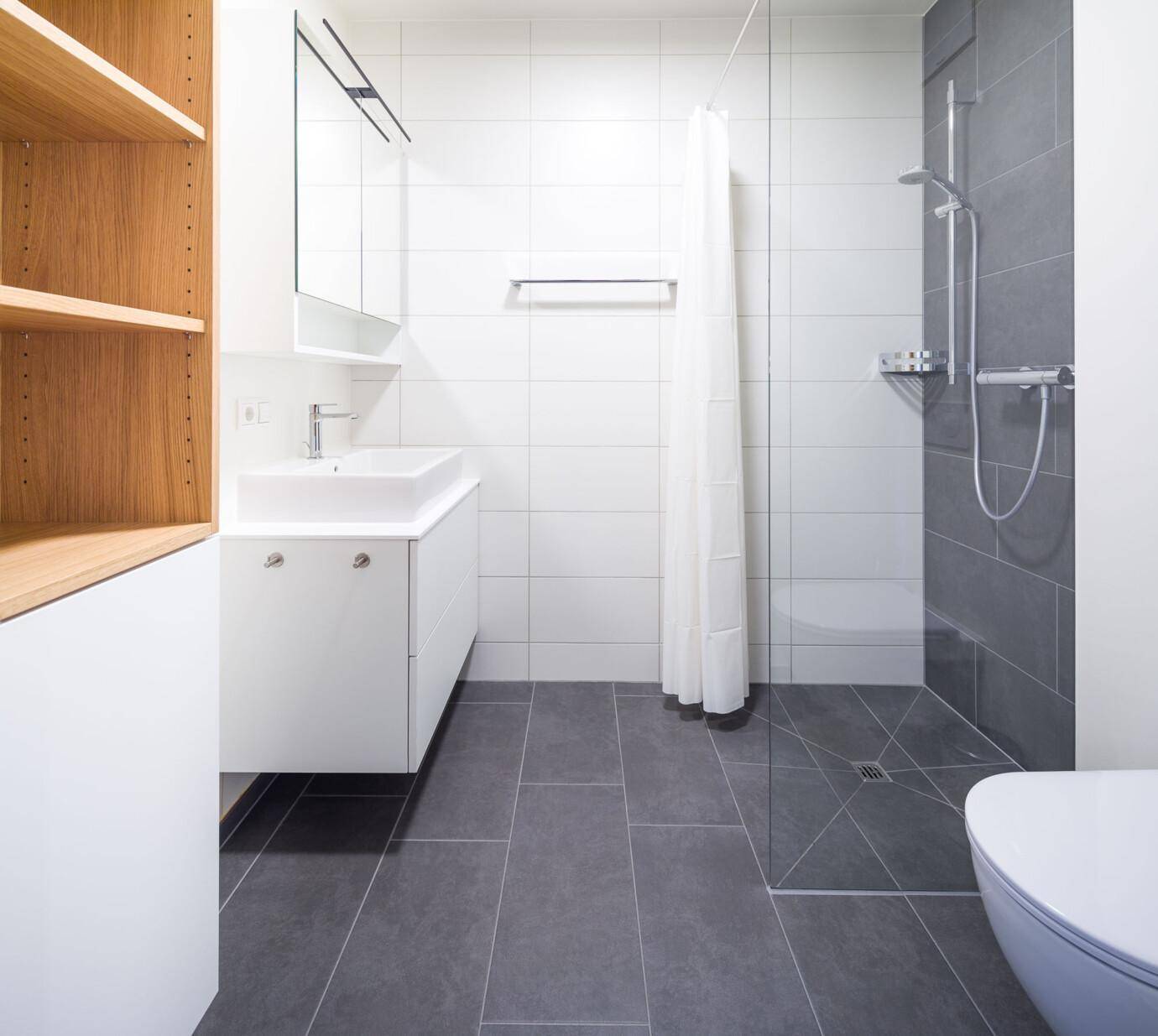 Ein modernes Badezimmer mit weiß gefliesten Wänden, einem Waschbeckenunterschrank, Duschbereich und dunklem Fliesenboden. Links ist ein Bücherregal aus Holz zu sehen (David).