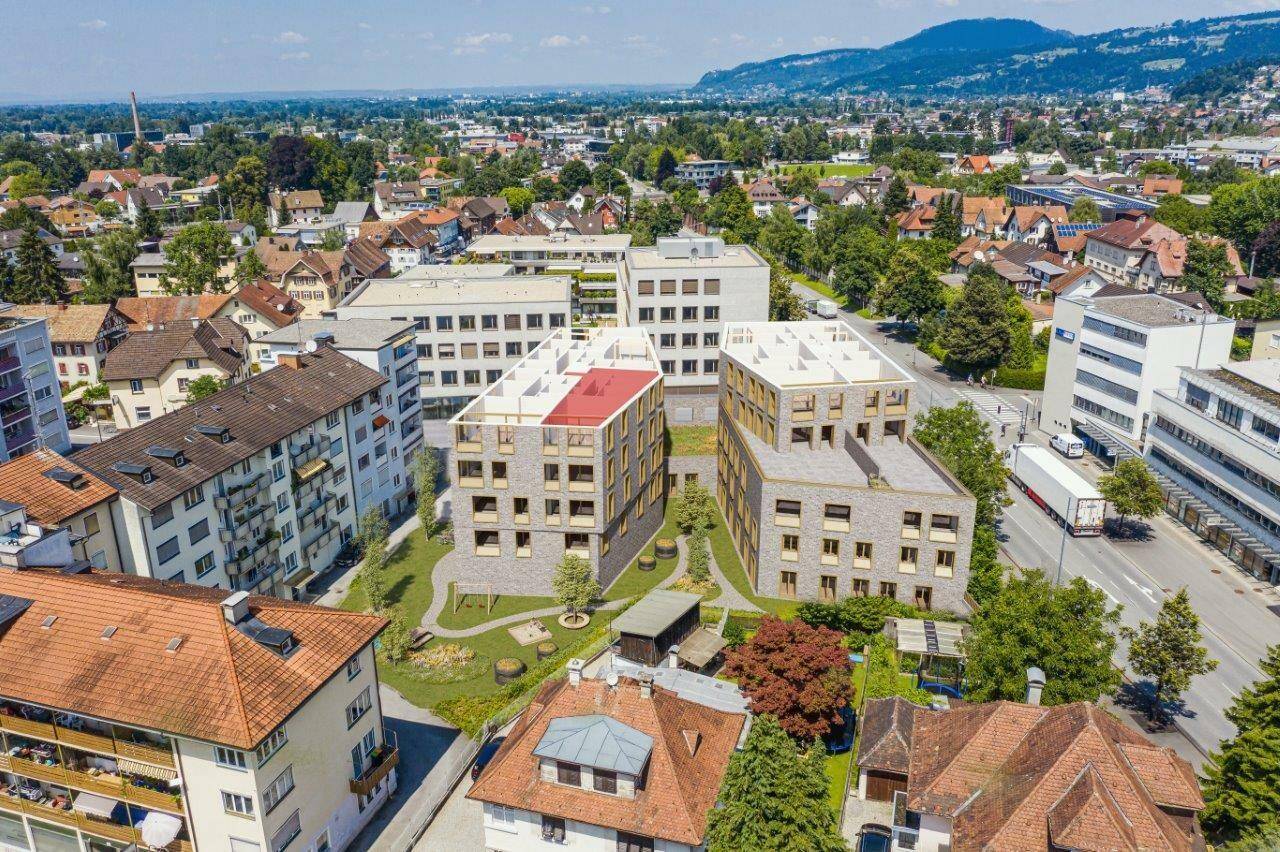 Luftaufnahme eines modernen Wohnkomplexes mit Grünflächen, umgeben von bestehenden Gebäuden und einer Straße, mit Bergen im Hintergrund.