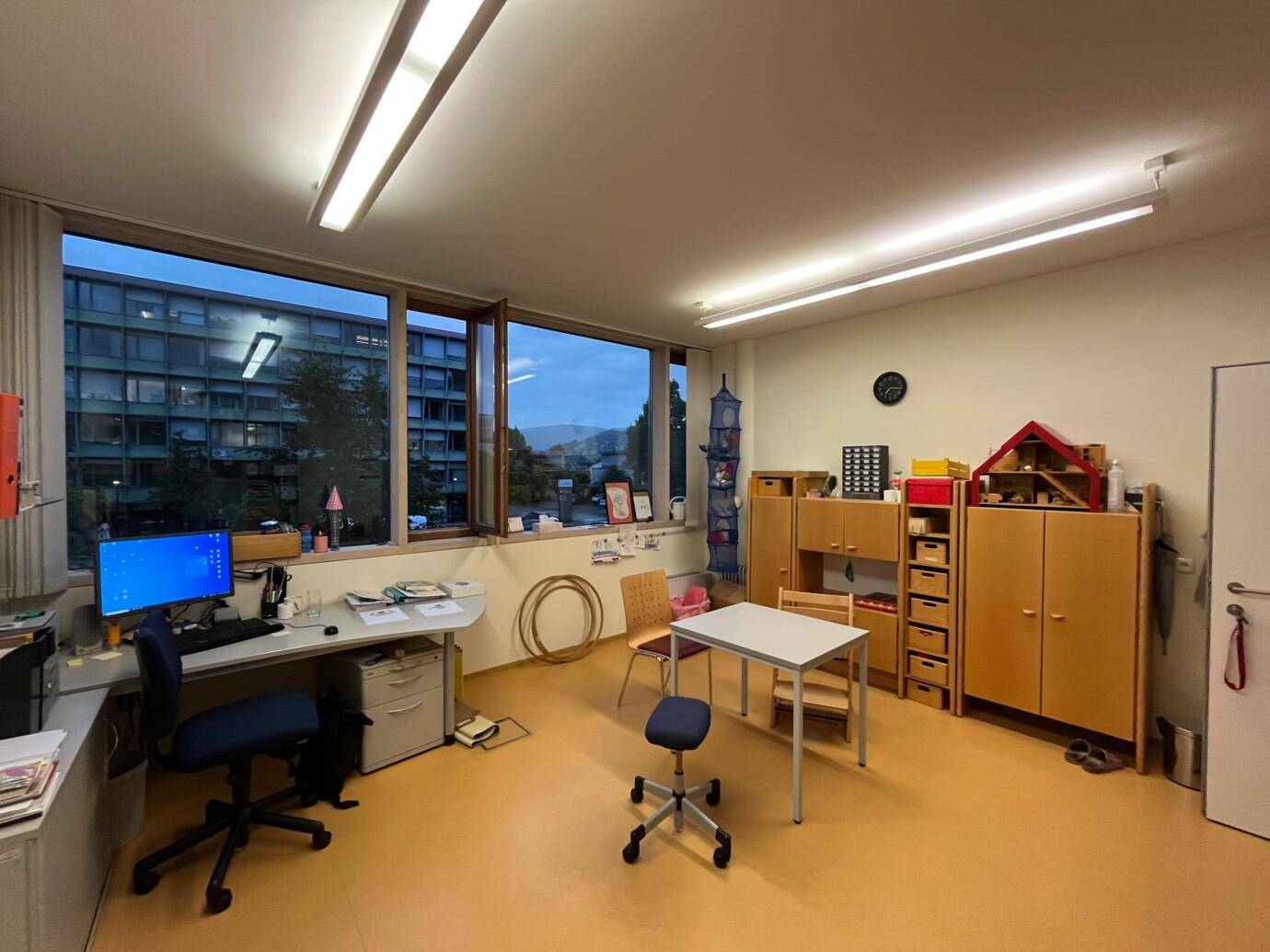 Büro- oder Therapieraum mit Schreibtisch, Stühlen, Holzmöbeln und großen Fenstern mit Stadtblick.