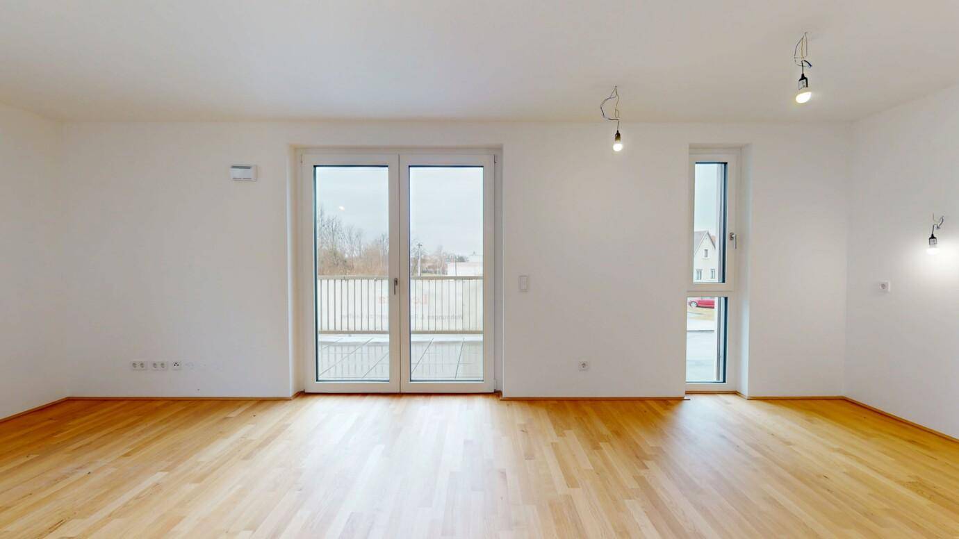 Geräumiges, leeres Wohnzimmer mit hellem Parkettboden und großzügigem Balkonzugang.