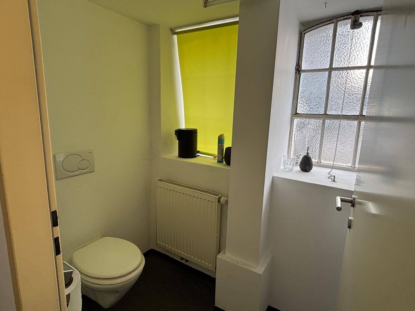 Kleine Toilette mit Wand-WC, Heizkörper und Fenster mit gelbem Rollo.