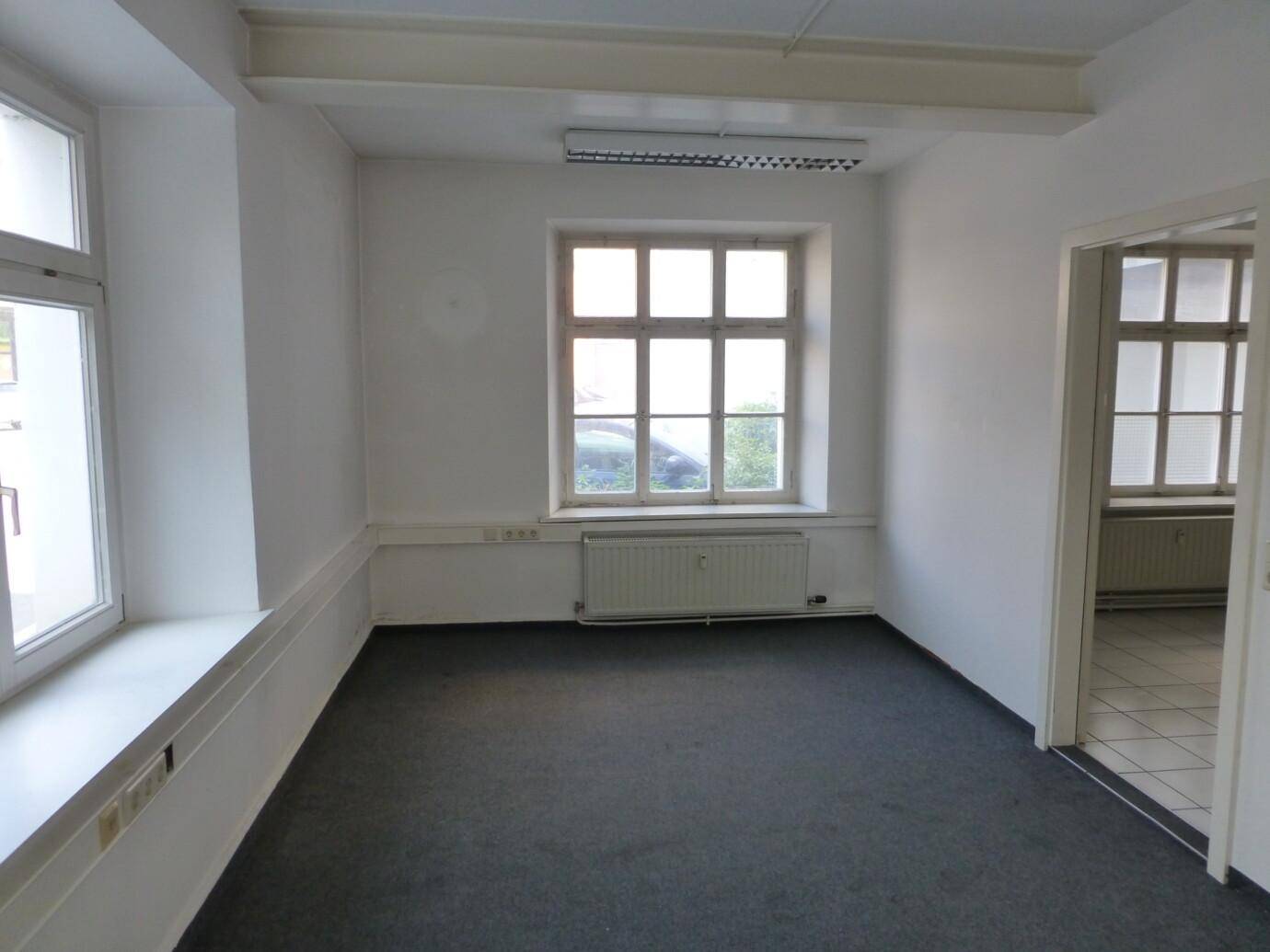 Heller Büroraum mit großen Fenstern, Teppichboden und sichtbaren Deckenbalken.