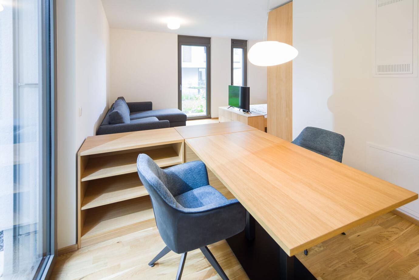 Moderner Wohn- und Essbereich mit Holztisch, blauen Stühlen, grauem Sofa und großen Fenstern zu einem Garten (David).