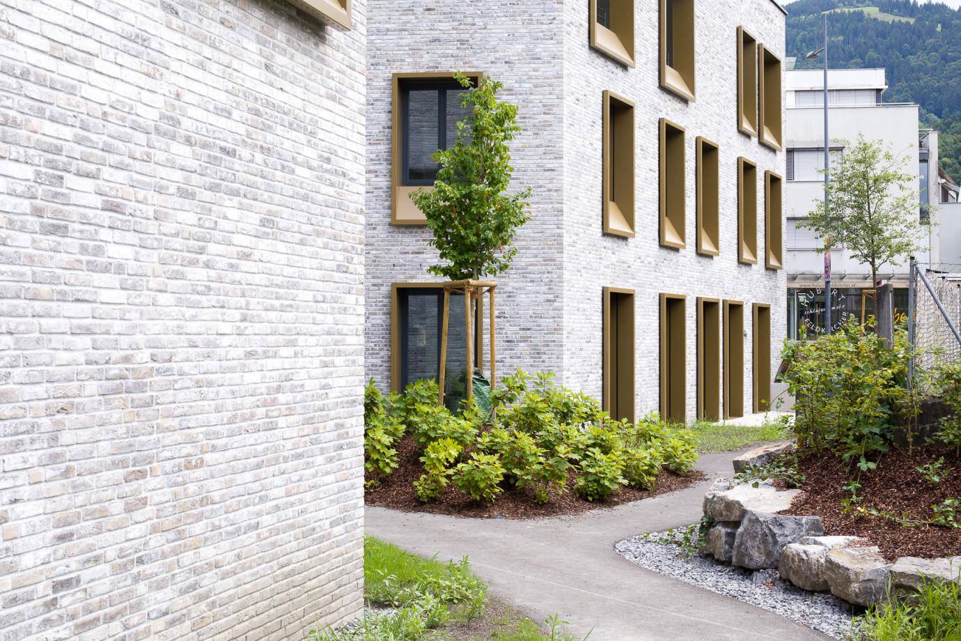 Moderne Gebäudefassade aus hellem Backstein mit markanten Fensterrahmen und begrüntem Außenbereich vor Bergkulisse.