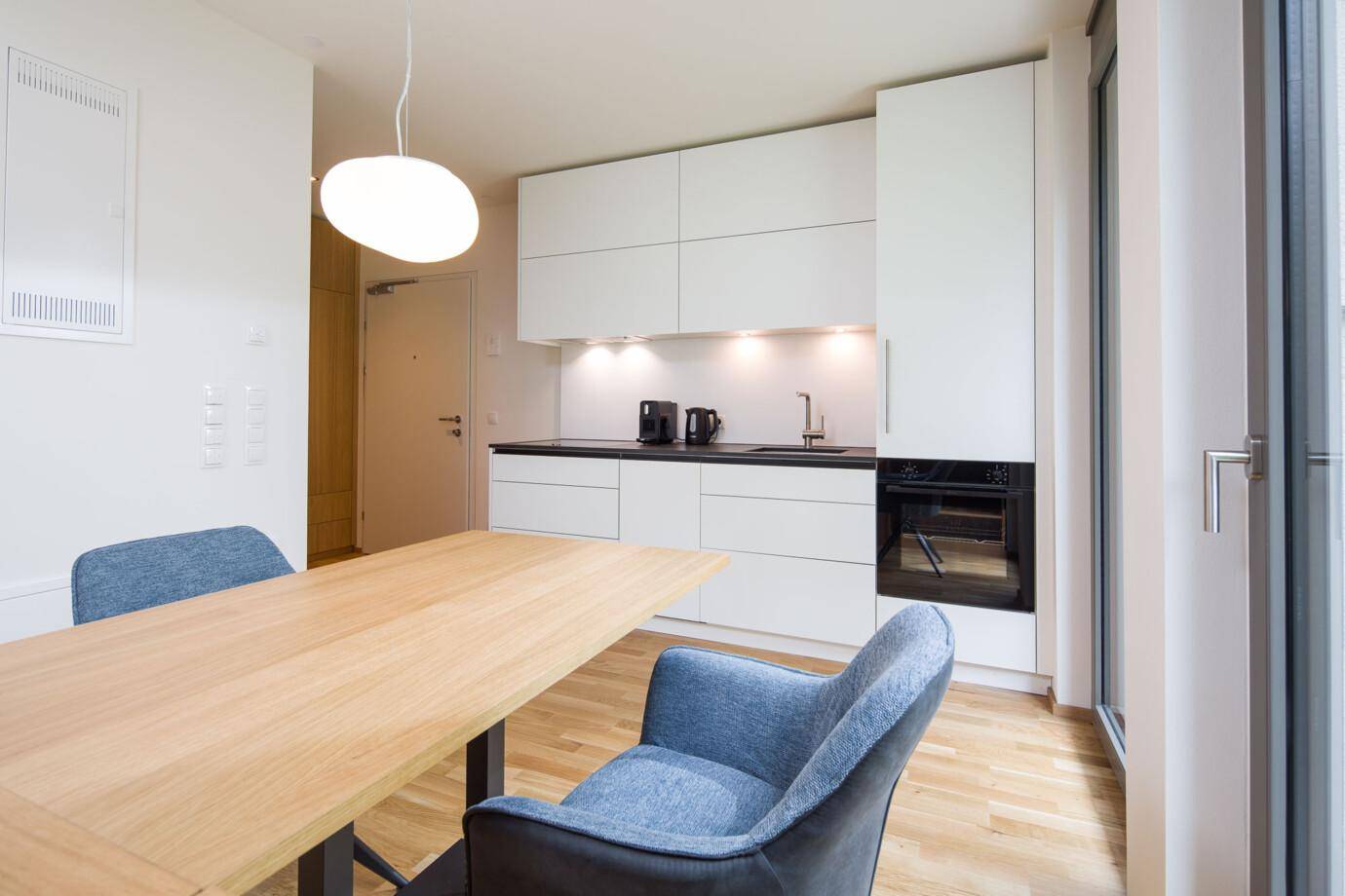 Moderne, offene Küche in einem Apartment mit weißer Einbauküche, Holztisch mit blauen Stühlen und Parkettboden (David).