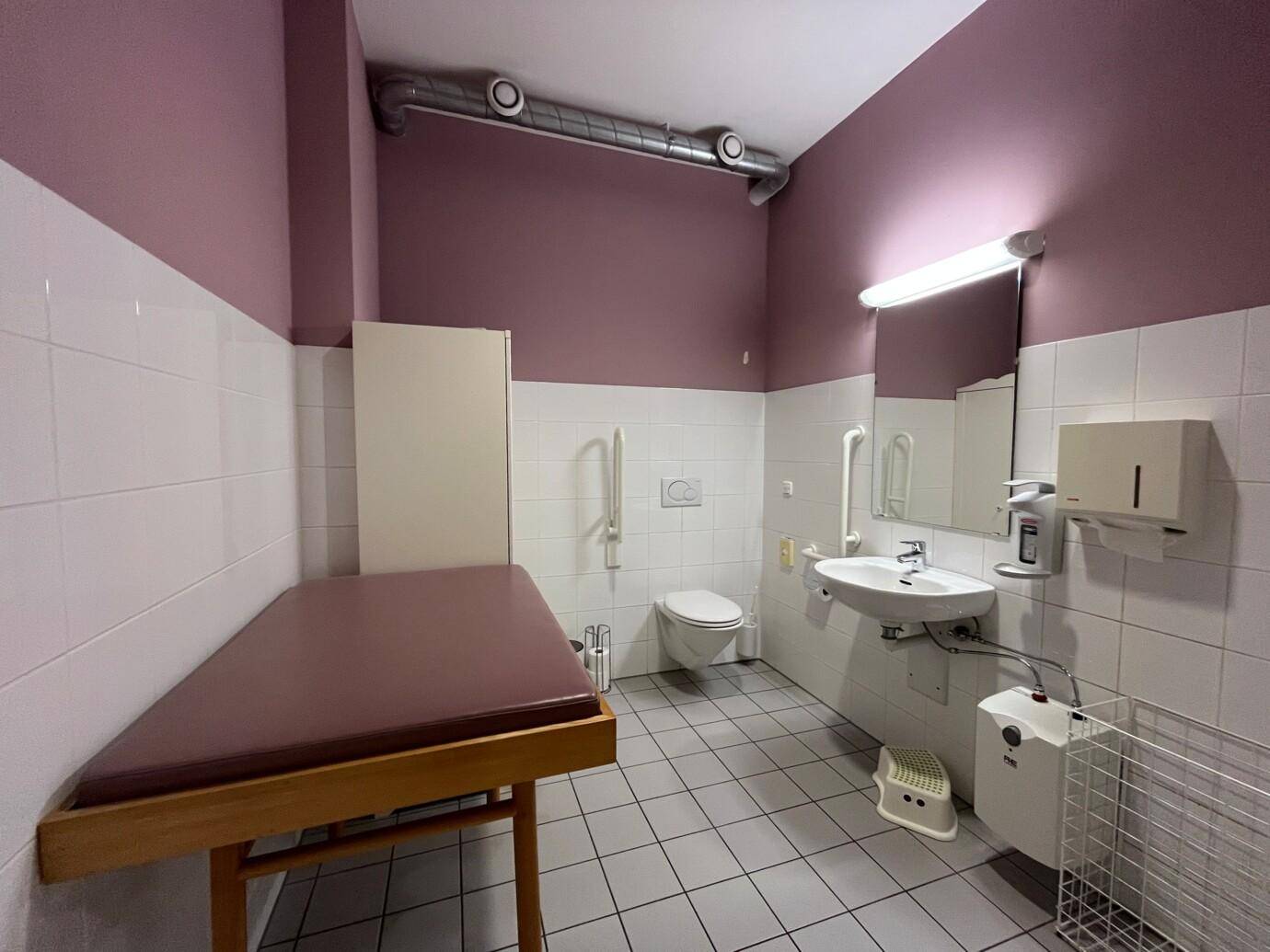Barrierefreies Badezimmer mit Wickeltisch, Toilette, Waschbecken und Haltegriffen an den Wänden.