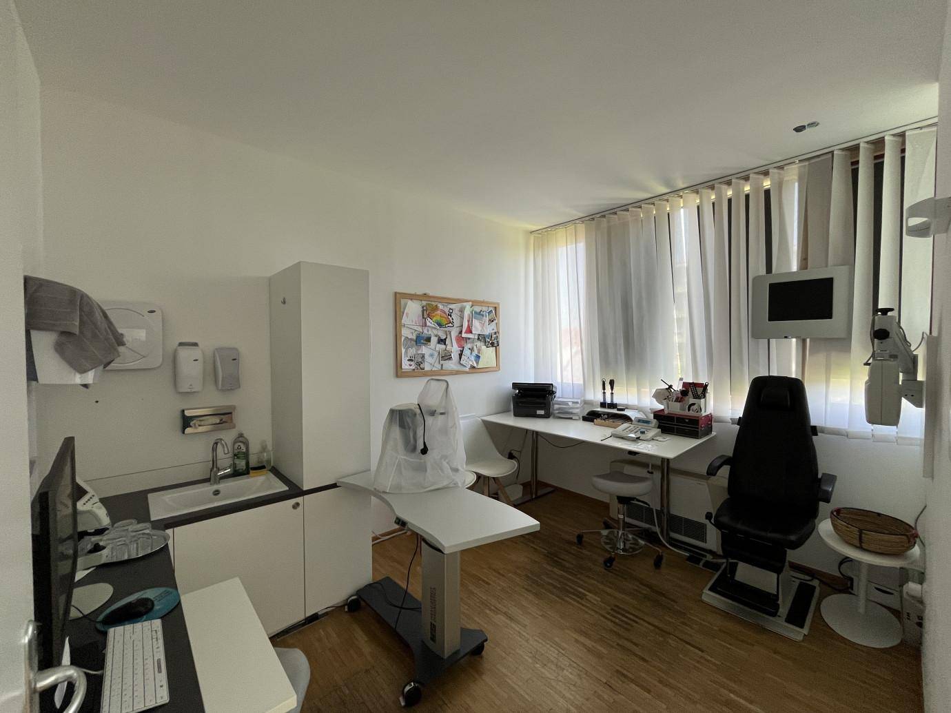 Praxisraum mit medizinischer Ausstattung, Schreibtisch, Waschbecken und Fenster mit Jalousien für Privatsphäre.