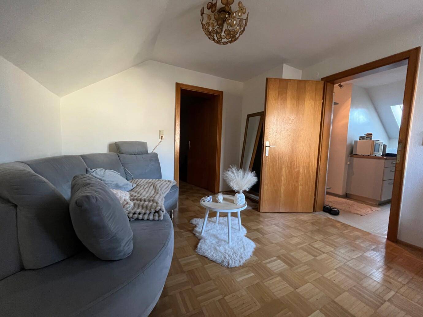 Ein Zimmer mit einer grauen Couch, einem runden Beistelltisch auf einem Kunstfellteppich und zwei offenstehenden Holztüren.