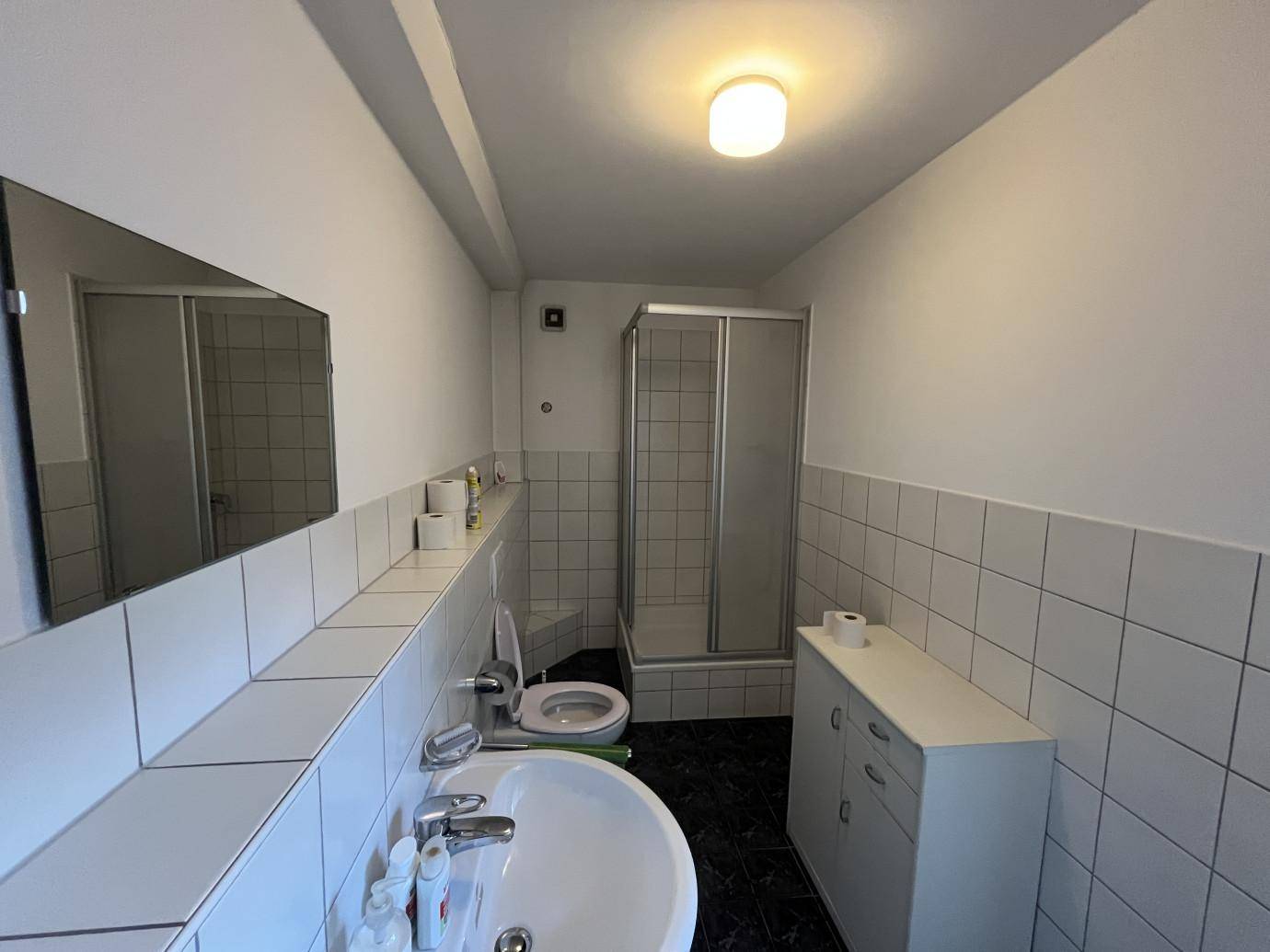 Funktionales Badezimmer mit weißen Fliesen, Waschbecken, Toilette und einer Duschkabine, gut beleuchtet.