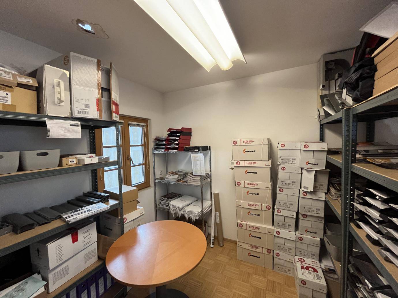 Lagerraum oder Archiv mit Regalen voller Kartons und Akten, einem kleinen Tisch und einem Fenster.
