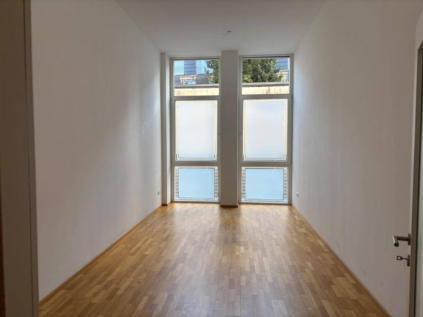 Geräumiger, leerer Raum mit Holzparkett und großen Fenstern, die für viel Helligkeit sorgen.
