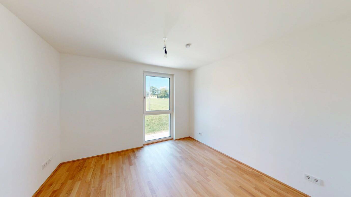 Helles Schlafzimmer mit Parkettboden und einem Fenster mit Blick ins Grüne.