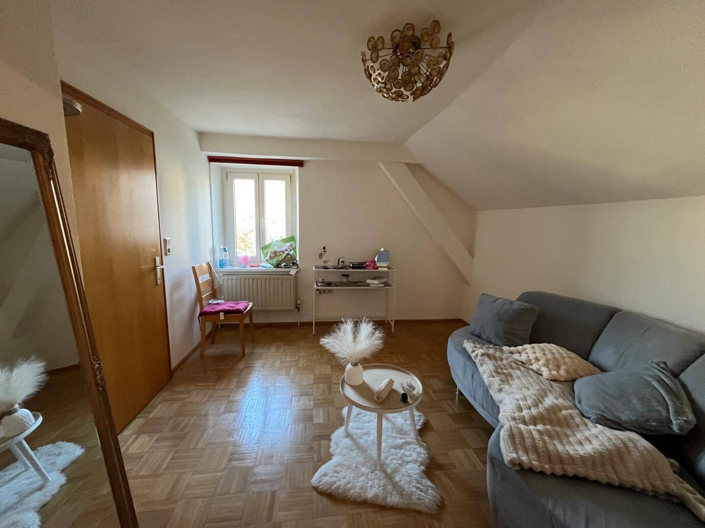 Wohnzimmer mit grauem Sofa und Decke, Beistelltisch auf Schaffell-Teppich, Holzboden, Fenster und Holztür.