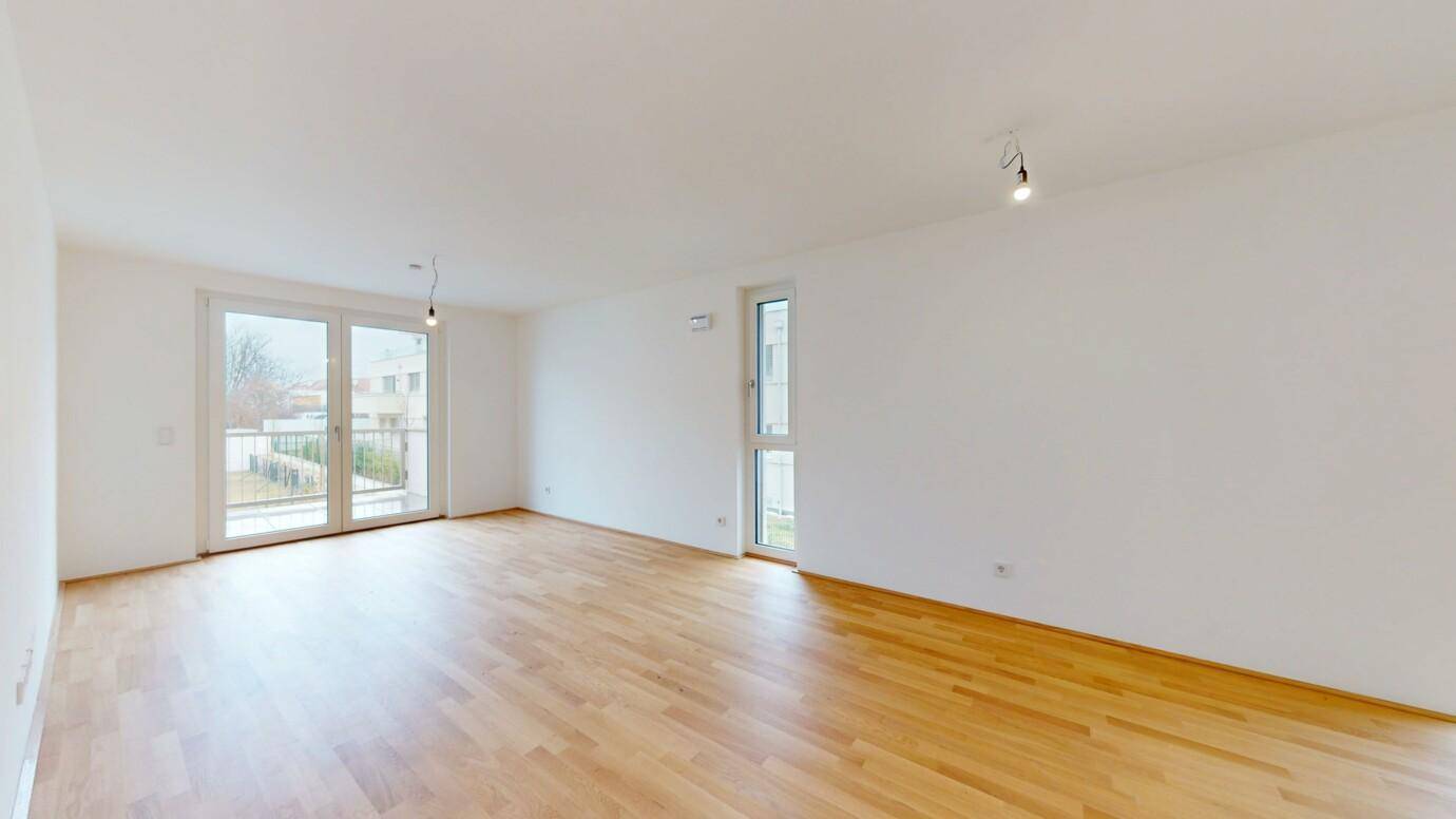 Helles, leeres Wohnzimmer mit Holzboden und Zugang zu einem Balkon.