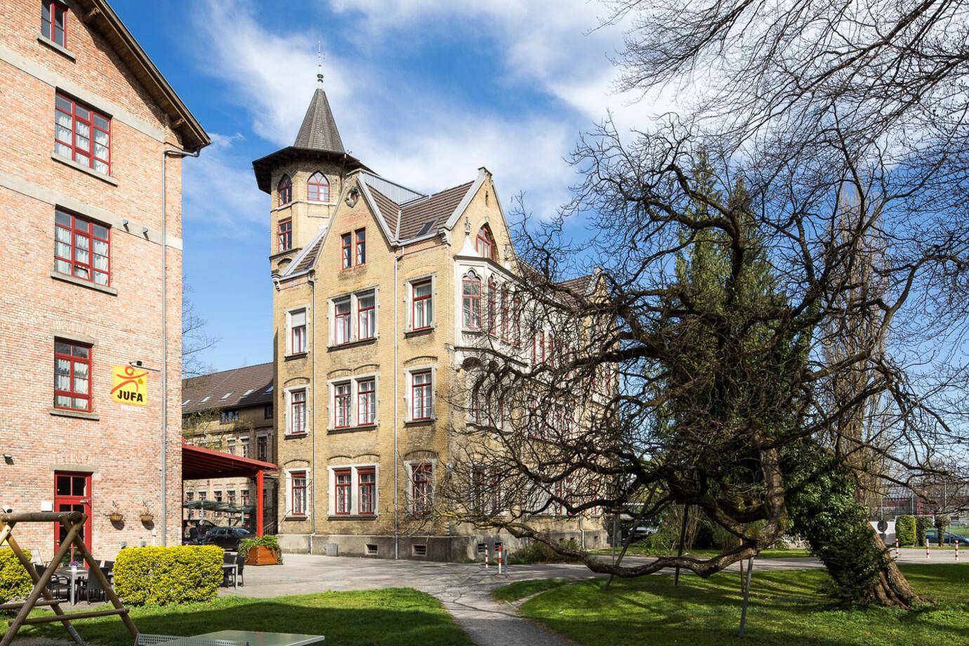 Blick auf das Benger Areal mit einem gelben Gebäude, grünen Wiesen und einem großen kahlen Baum im Vordergrund.
