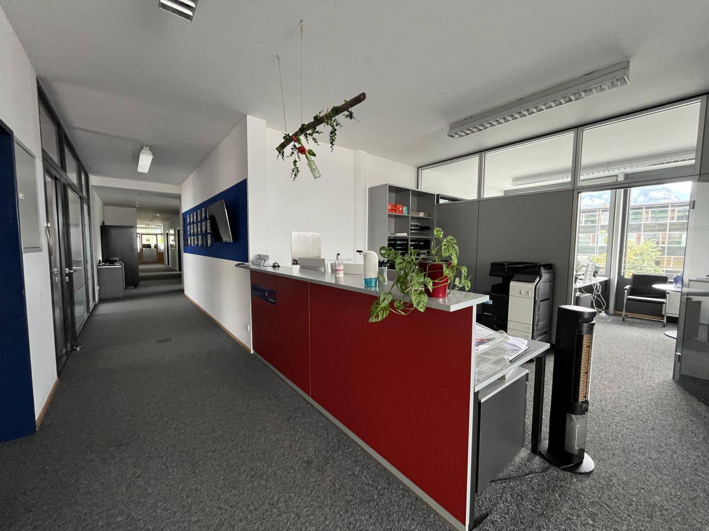 Empfangsbereich mit roter Theke, Bürozellen und einem langen Flur, der zu weiteren Räumen führt.