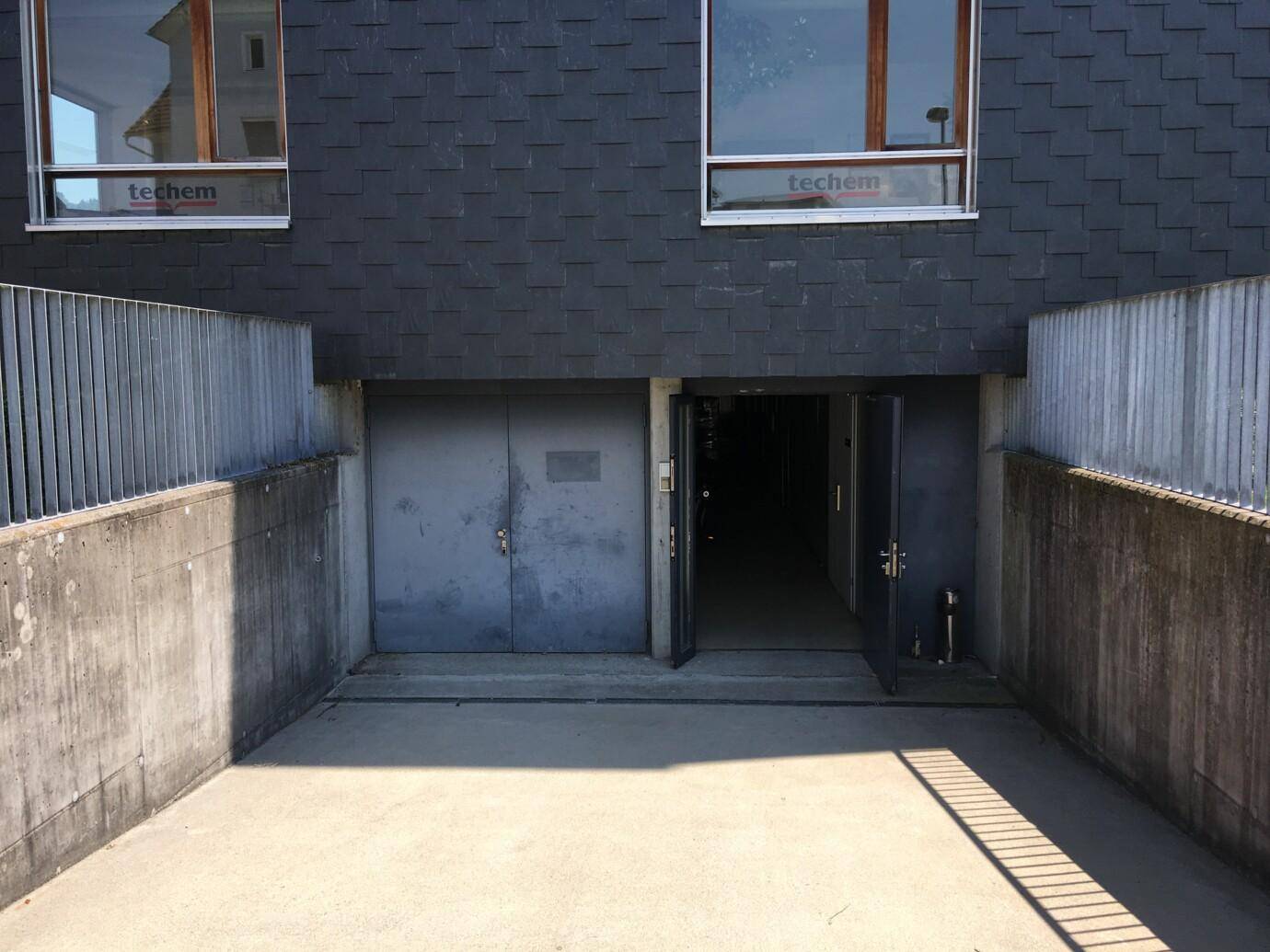 Eingangsbereich zu einer Tiefgarage oder einem Kellergeschoss mit Betonwänden, Metallgeländer und dunkler Gebäudefassade.