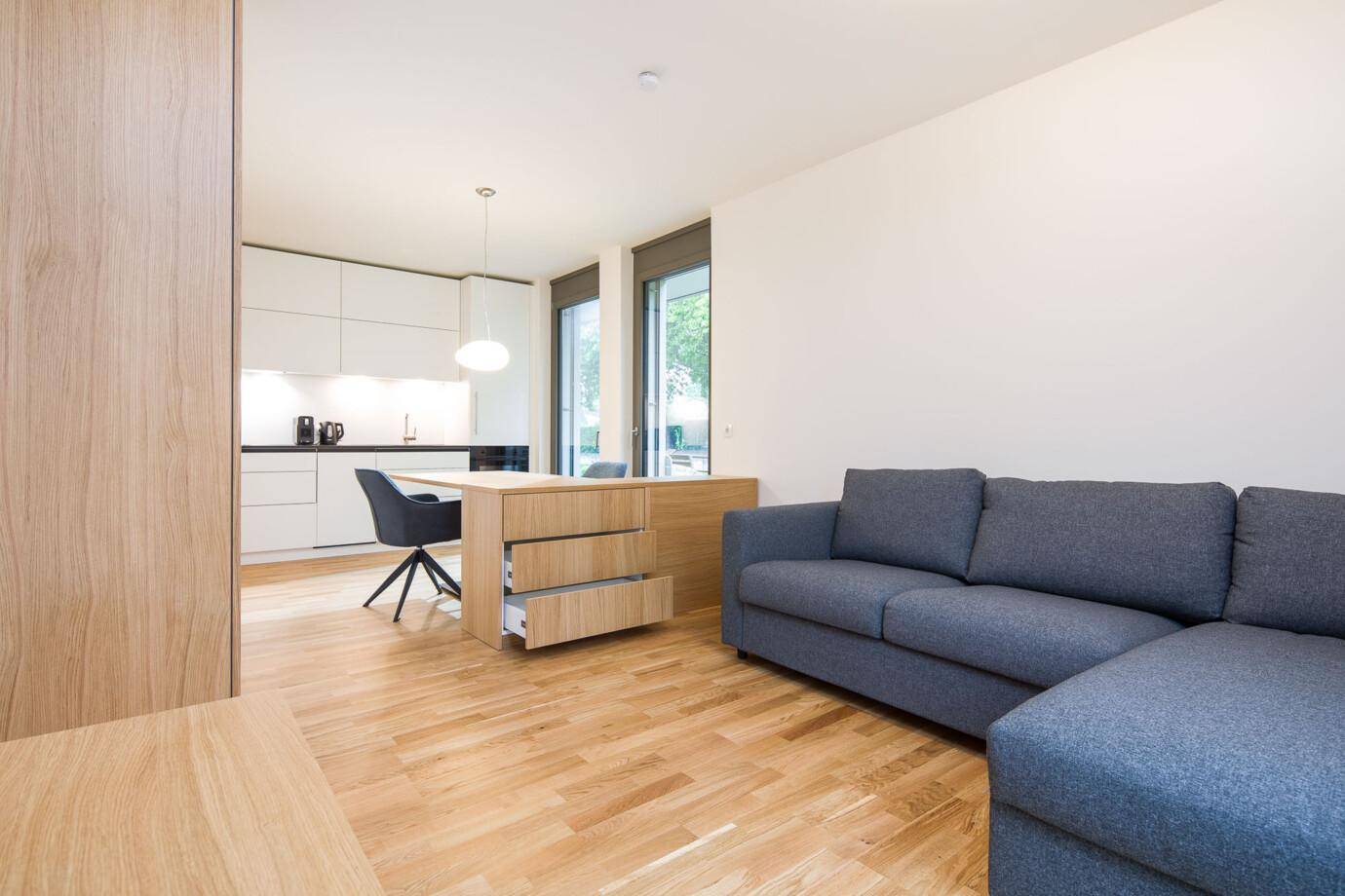 Moderner offener Wohn- und Essbereich mit grauer Couch, Holzboden und weißer Hochglanzküche. Ein Esstisch mit offenen Schubladen steht am Übergang (David).