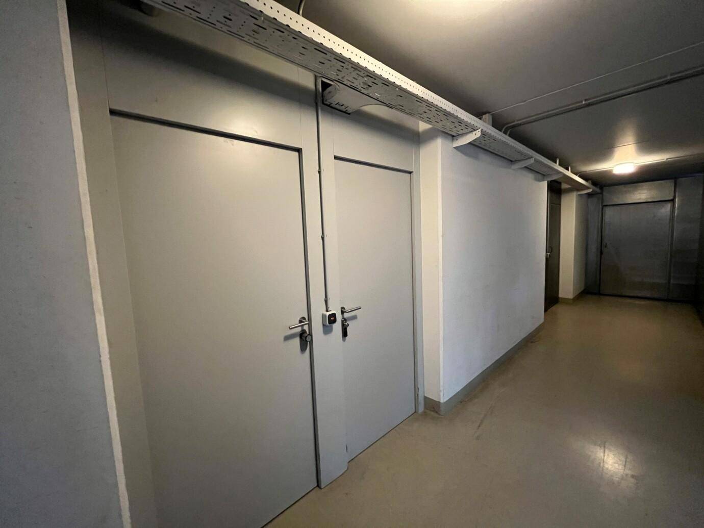Innenansicht eines Kellerflurs mit zwei grauen Türen, weißen Wänden und sichtbaren Kabeltrassen an der Decke.