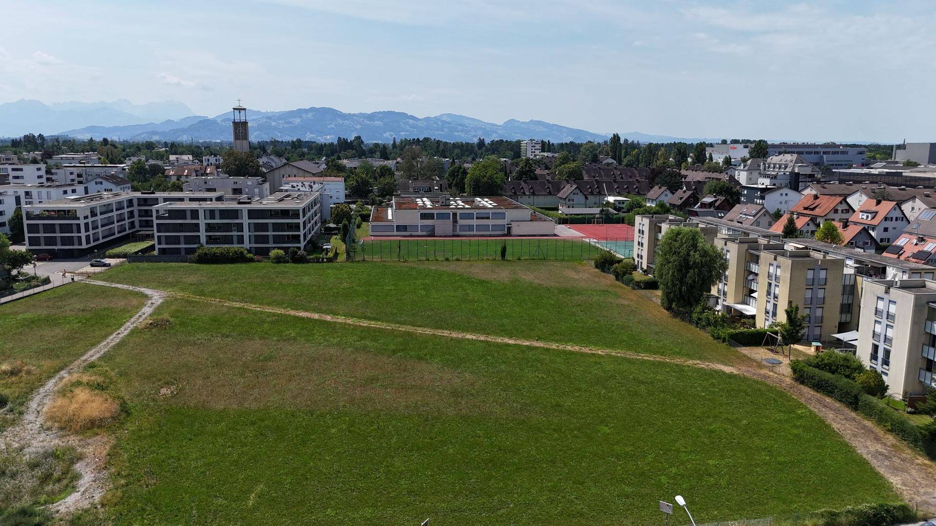 Luftaufnahme eines großen Grundstücks mit umliegenden Wohngebäuden, Sportanlagen und Bergblick.