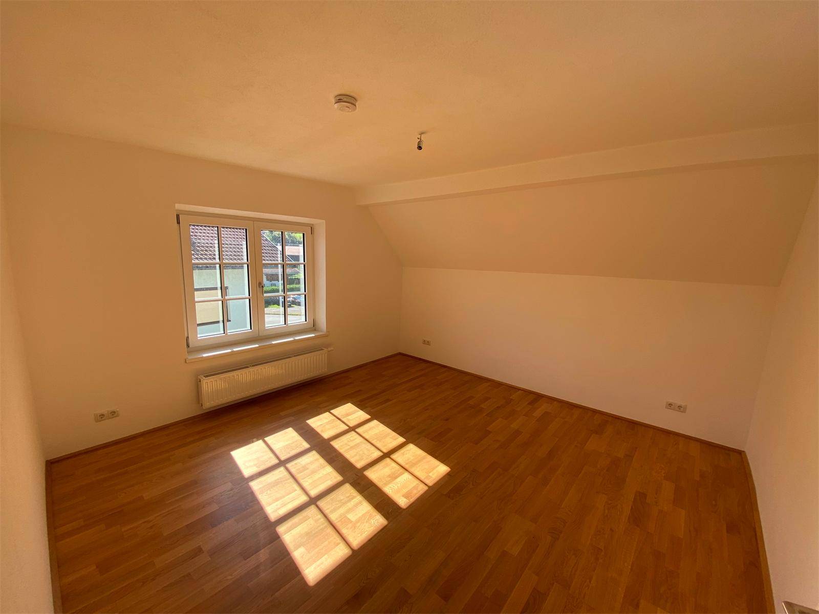 Sonniges Zimmer im Dachgeschoss mit Parkettboden, Fenster und sichtbaren Dachschrägen.
