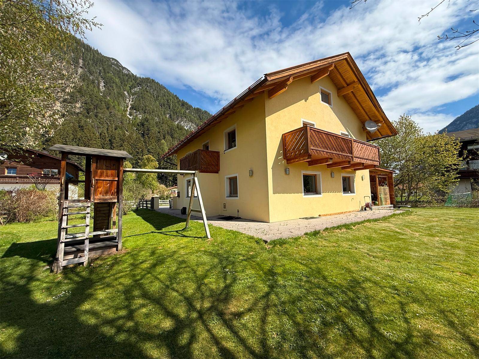 Gelbes Wohnhaus mit Holzbalkonen und einem Spielplatz im grünen Garten vor beeindruckender Bergkulisse.