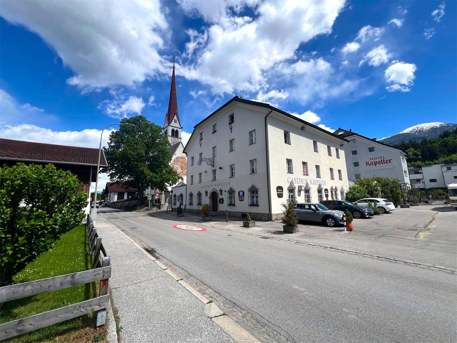 Historisches Gasthofgebäude an einer Straße mit parkenden Autos und Bergen im Hintergrund.
