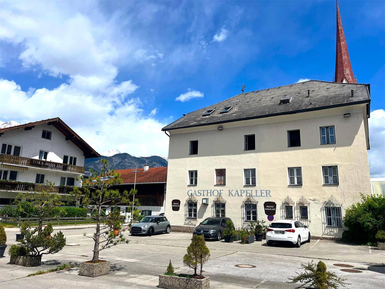 Ansicht des Gasthofs mit Parkplatz und Bergen im Hintergrund unter einem bewölkten Himmel.