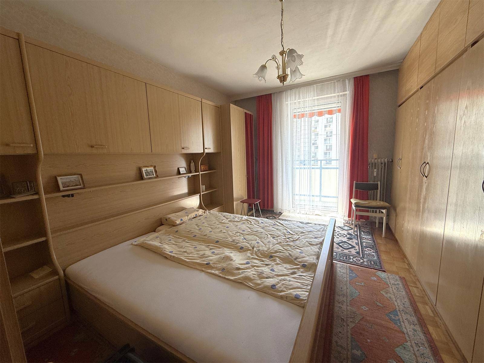 Schlafzimmer mit Holzmöbeln, Fenster mit roten Vorhängen und Blick auf die Umgebung.