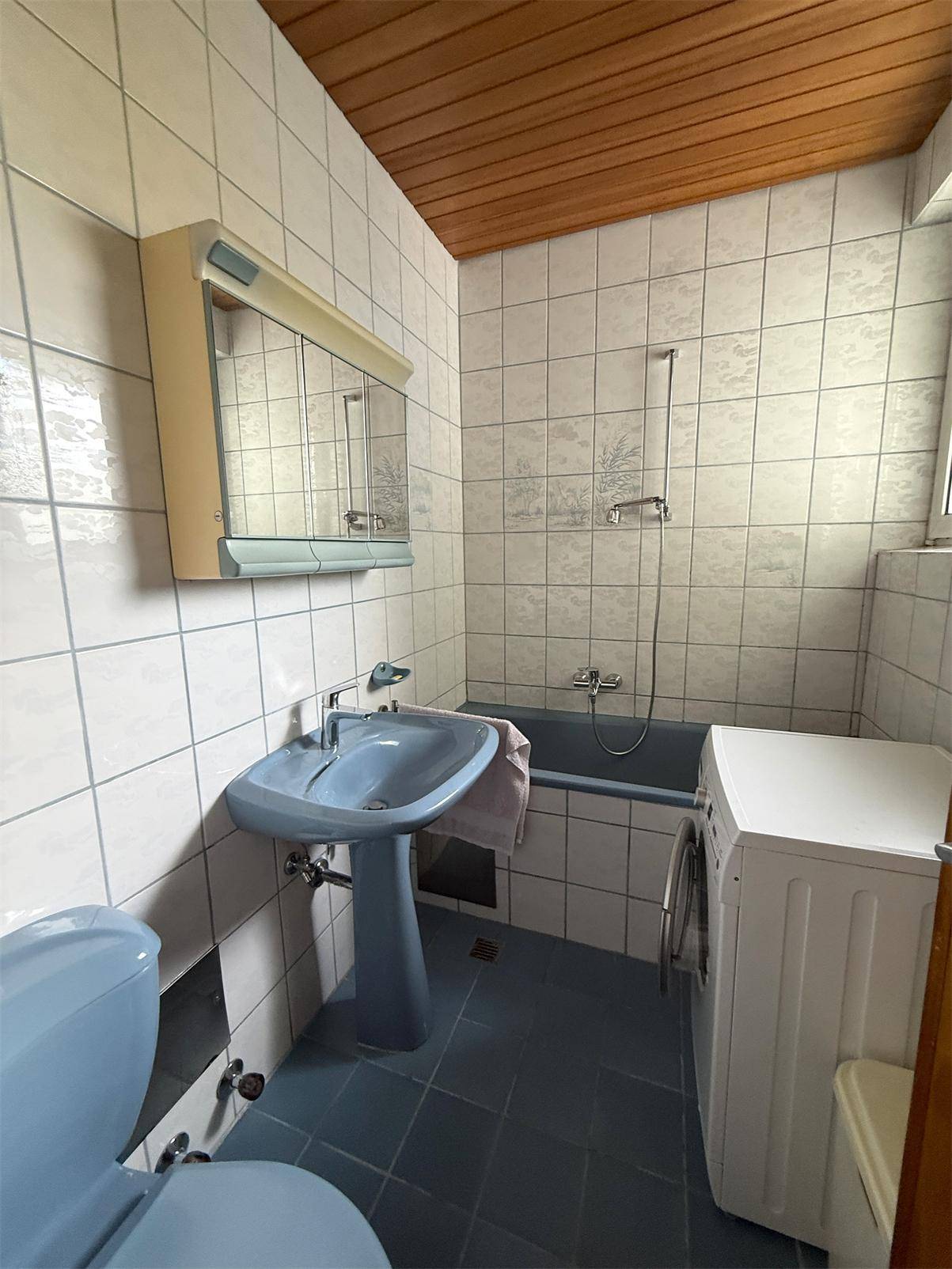 Badezimmer mit blauen Sanitärobjekten, weißen Fliesen, Holzdecke und einer Waschmaschine.