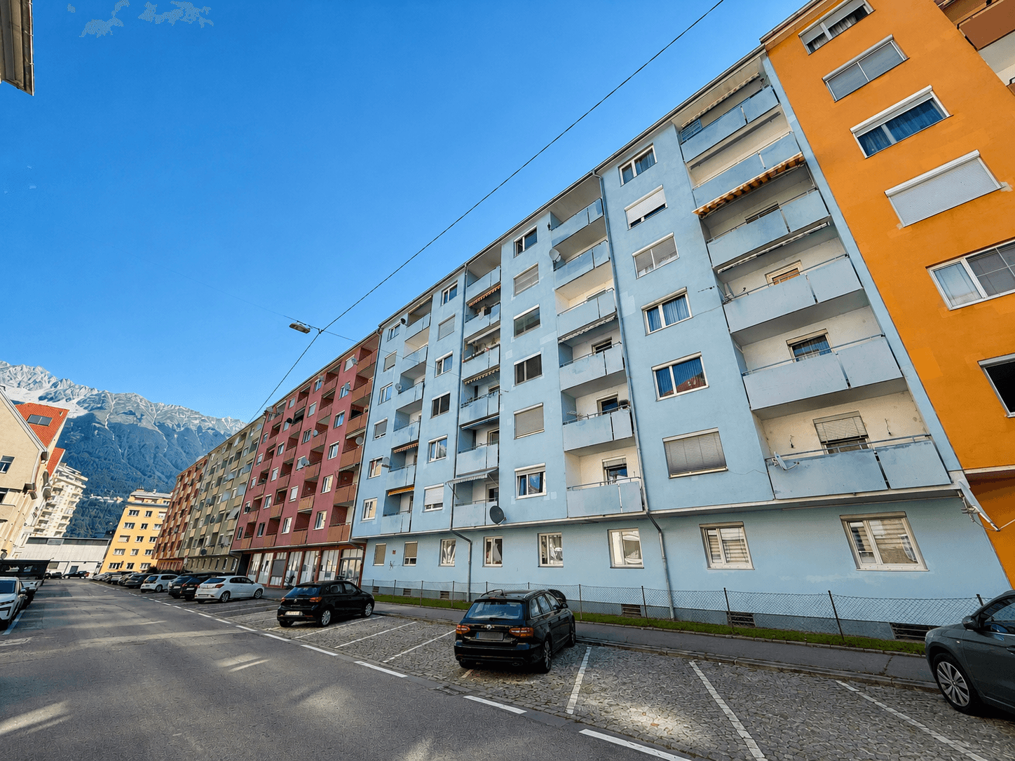 Wohngebäude entlang einer Straße mit geparkten Fahrzeugen und den Bergen im Hintergrund.