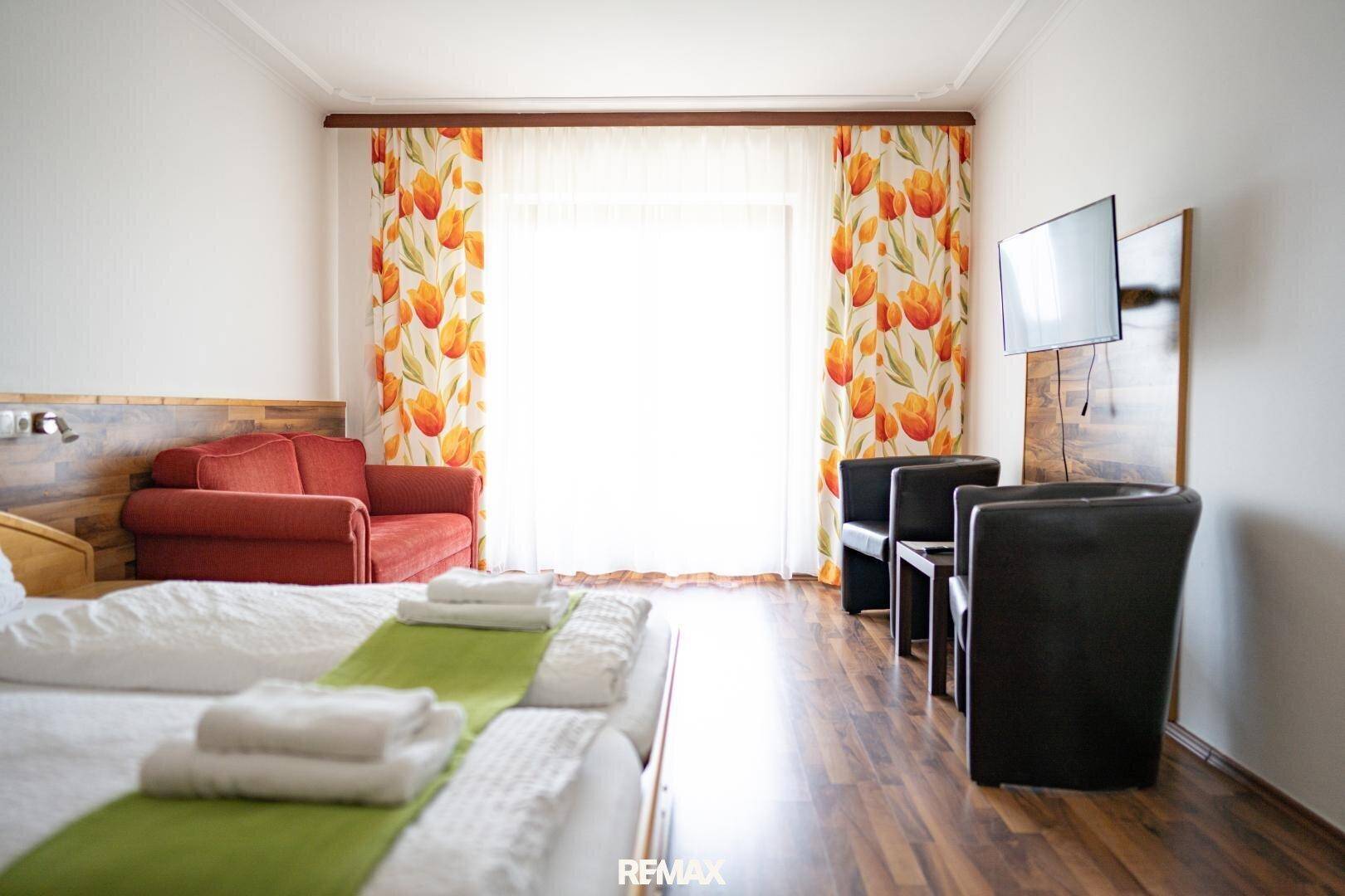 Helles Schlafzimmer mit Bett, Sofa, Fernseher und großem Fenster, das viel Licht spendet.