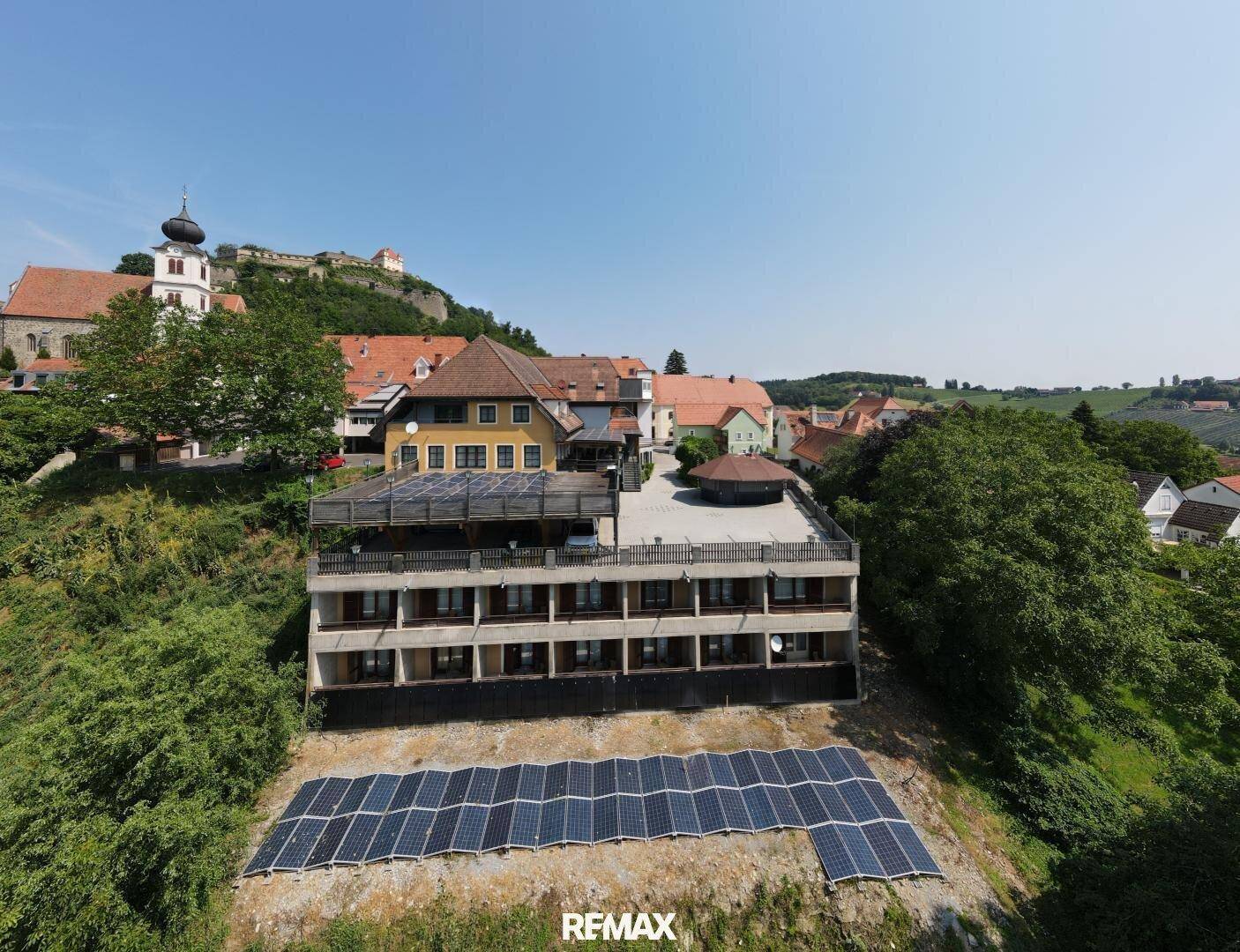 Luftaufnahme eines modernen Gebäudes mit großer Terrasse und Photovoltaikanlage auf dem Hang, umgeben von grüner Landschaft.