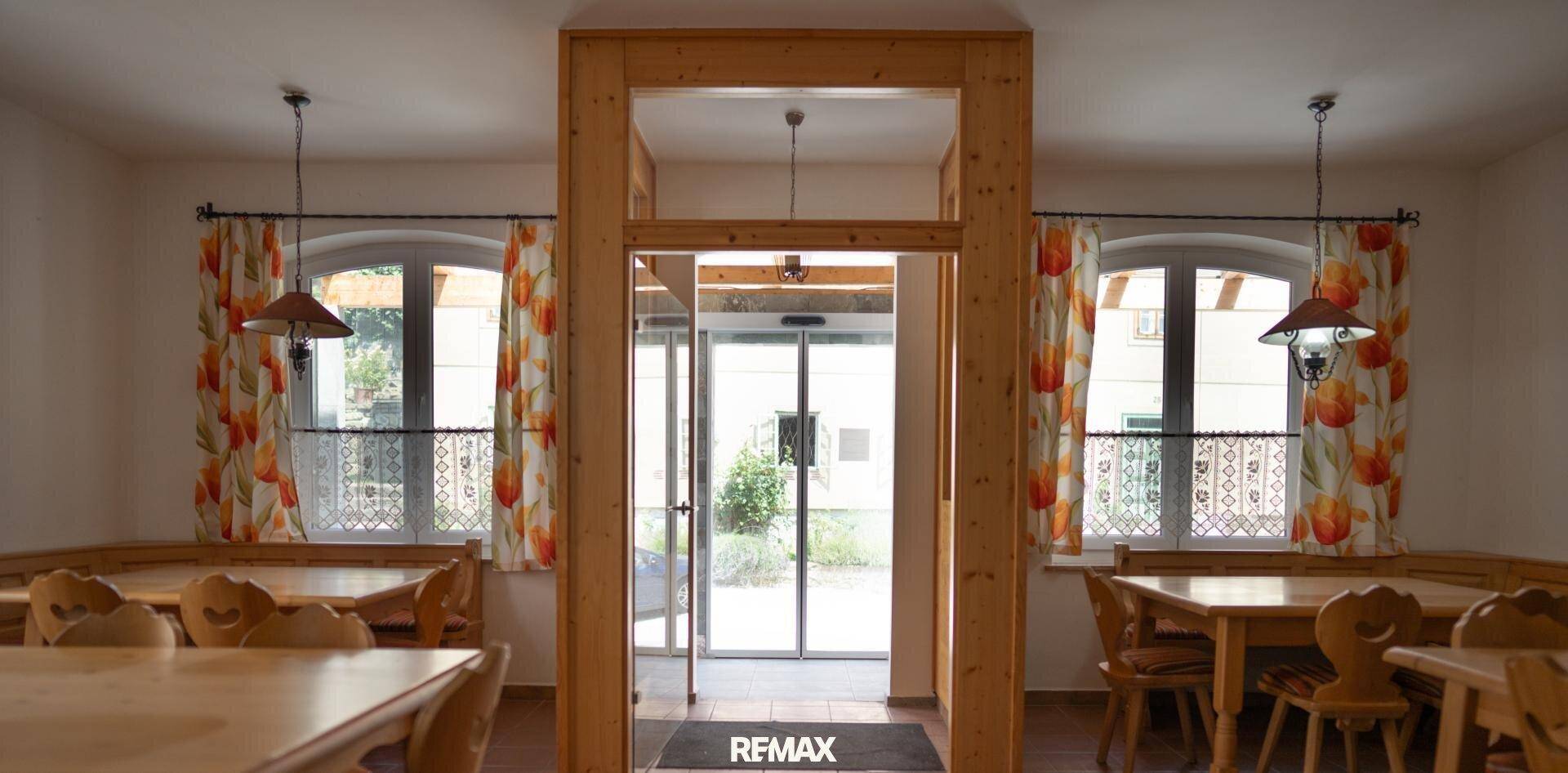 Gemütlicher Speisesaal mit traditionellen Holztischen und Bänken, hell durch Fenster mit Vorhängen.